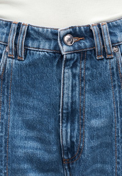 Κοντινό πλάνο στη ζώνη μέσης από μπλε τζιν με μεταλλικό κουμπί και εμφανείς θηλιές ζώνης, που δείχνει τις λεπτομερείς ραφές και την υφή του υφάσματος.
