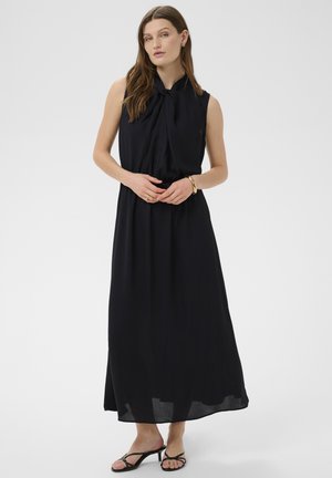 Kobieta o długich brązowych włosach ubrana w czarną długą sukienkę bez rękawów z kokardą przy szyi oraz czarne sandały na obcasie z paskami.