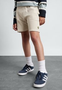 Beige Fleece-Shorts mit einem kleinen gelben gestickten Logo, kombiniert mit Navy-Sneakern und weißen Socken, vor einem grauen Hintergrund.