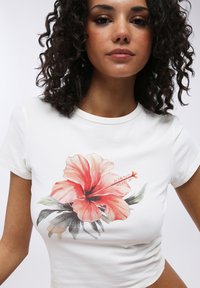 T-shirt in cotone bianco con una grande stampa di un fiore di ibisco colorato accompagnato da foglie verdi, design aderente e maniche corte.