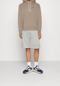 Sweat à capuche marron avec cordons, short gris et baskets bleu marine. La tenue présente une coupe décontractée et des textures de tissu lisses.