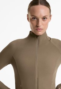 Långärmad, åtsittande jacka med dragkedja i en dämpad beige-brun färg, med ribbad textur, hög krage och två sidofickor.