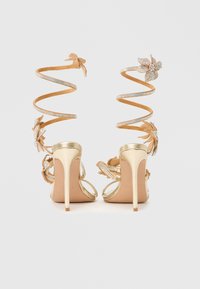 Kuldsete kõrgete kontsadega sandaalide rihmad on spiraalsed ja kaunistatud rhinestone'ide ning lillemotiividega, pehme seemisnahkse tekstuuriga ning läbipaistva esiosaga.