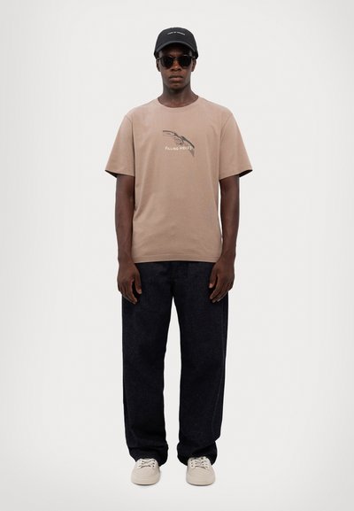 Beige Baumwoll-T-Shirt mit grafischem Druck, dunkle Jeanshose, weiße Schuhe, schwarze Cap und Sonnenbrille, vor einem neutralen Hintergrund stehend.