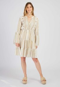 Weißes Kleid mit goldenen geometrischen Mustern, V-Ausschnitt, schwingenden langen Ärmeln und gestuftem Rüschensaum. Kombiniert mit beigen Plateausandaletten.