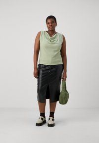 Blusa sin mangas de color verde claro con escote drapeado, falda midi de cuero negra con abertura frontal, bolso acolchado verde, zapatos crema.