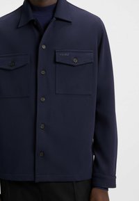Camicia blu navy a maniche lunghe con tessuto testurizzato, due tasche sul petto e un logo discreto sulla tasca sinistra.