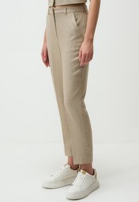 Beige maßgeschneiderte Hose mit strukturiertem Muster, seitlichen Taschen und schmal zulaufendem Bein, kombiniert mit weißen Sneakern.