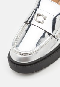 Mocassin en argent brillant avec une épaisse semelle en caoutchouc noir et un emblème métallique "C" sur la sangle au-dessus de la chaussure.