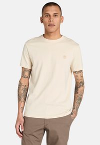 Béžové bavlněné tričko s kulatým výstřihem a krátkými rukávy; na hrudi má malé tónované logo. Hladká textura a přiléhavý střih.