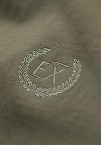 Bestickter weißer Lorbeerkranz, der die Buchstaben "EX" auf olivgrünem Stoff umgibt.
