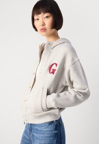 Noor naine lühikeste tumedate juustega, seljas heledast hallist kapuutsiga jakk punase "G" logoga, käed taskutes, koos siniste teksadega valge tausta ees.