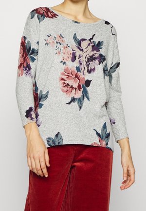 Vrouw die een grijze trui draagt met grote roze en paarse bloemenprints en rode corduroy broek, staand tegen een effen achtergrond.