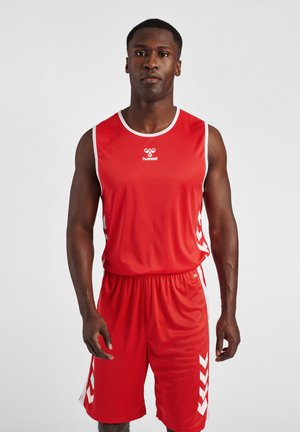 Atletisk mand står, iført en rød ærmeløs sportsjersey og matchende shorts med hvide chevron-mønstre på siderne.