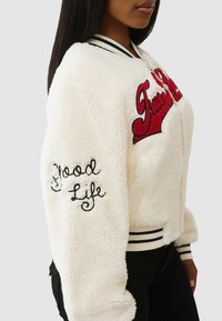 Biała kurtka bomber z futra ekologicznego z czerwonym wyhaftowanym napisem "True" oraz czarnym napisem "Good Life" na rękawie. Posiada prążkowane mankiety i kołnierz w paski.