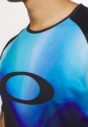 Mężczyzna ubrany w niebiesko-czarną koszulkę z gradientem z dużym czarnym owalnym logo z przodu, pokazany od szyi do połowy tułowia.