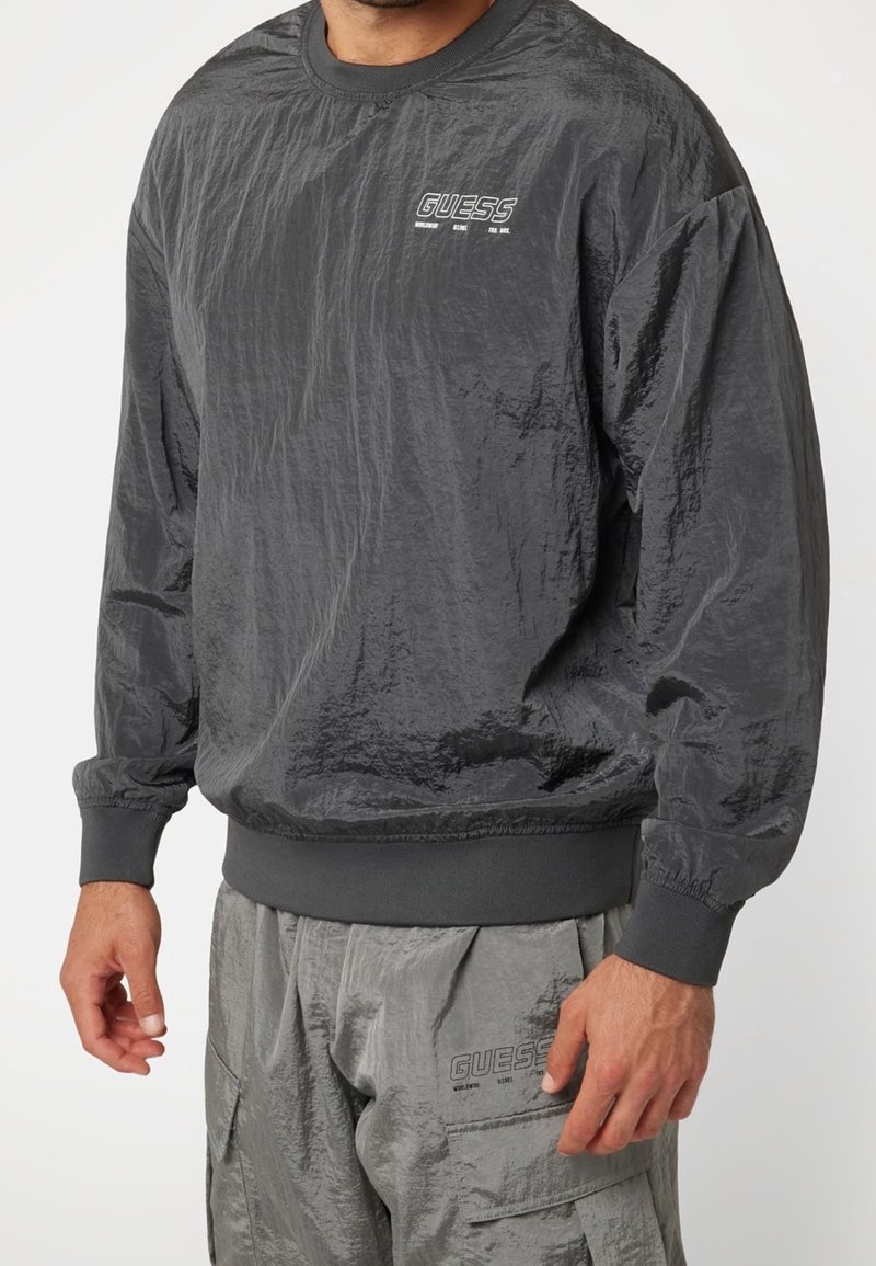 Homme portant un sweat-shirt gris foncé froissé à manches longues et un pantalon assorti, tous deux avec de petits logos blancs "GUESS" sur la poitrine et la cuisse.