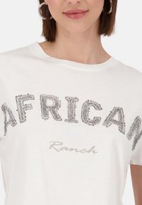 Weißes T-Shirt mit "AFRICAN" aus strukturierten silbernen Perlen und "Ranch" in hellgrau darunter. Weicher Stoff, lockere Passform, kurze Ärmel.