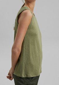Femme portant un haut en maille sans manches vert olive et un pantalon vert foncé, vue de côté sur fond gris uni.