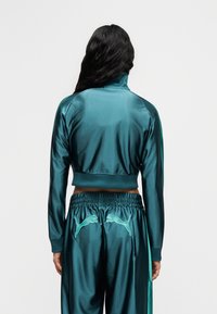 Άτομο με μακριά σκουρόχρωμα μαλλιά που φοράει σατέν φόρμα σε τιρκουάζ απόχρωση με δύο λογότυπα Puma στην πλάτη του παντελονιού, στέκεται μπροστά σε απλό φόντο.