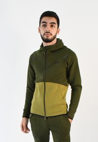 Sudadera con cremallera verde de diseño bicolor, más oscura en la parte superior y más clara en la parte inferior. Cuenta con un alto cuello y bolsillos laterales con cremallera. Tejido suave y texturizado.