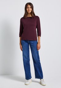 Frau, die vor einem schlichten Hintergrund steht und ein burgunderfarbenes Longsleeve, blaue gerade geschnittene Jeans und weiße Turnschuhe trägt.