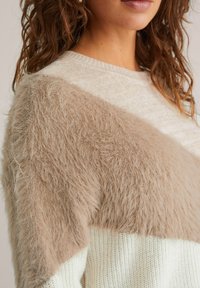 Pull en maille avec une superposition texturée marron duveteuse sur la partie supérieure, présentant une section inférieure crème douce et un col arrondi.