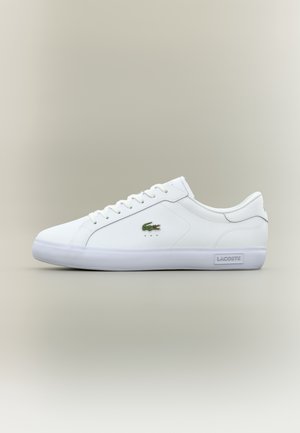 Hvid lavtop Lacoste sneaker med grønt krokodillelogo på siden og Lacoste-branding på hvid sål, fotograferet mod en neutral baggrund.