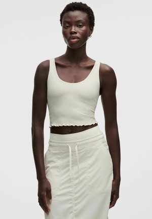 Kobieta z krótkimi kręconymi włosami, ubrana w białą prążkowaną, bezrękawną bluzkę typu crop top oraz białą spódnicę ze sznurkiem, stoi na jednolitym tle.