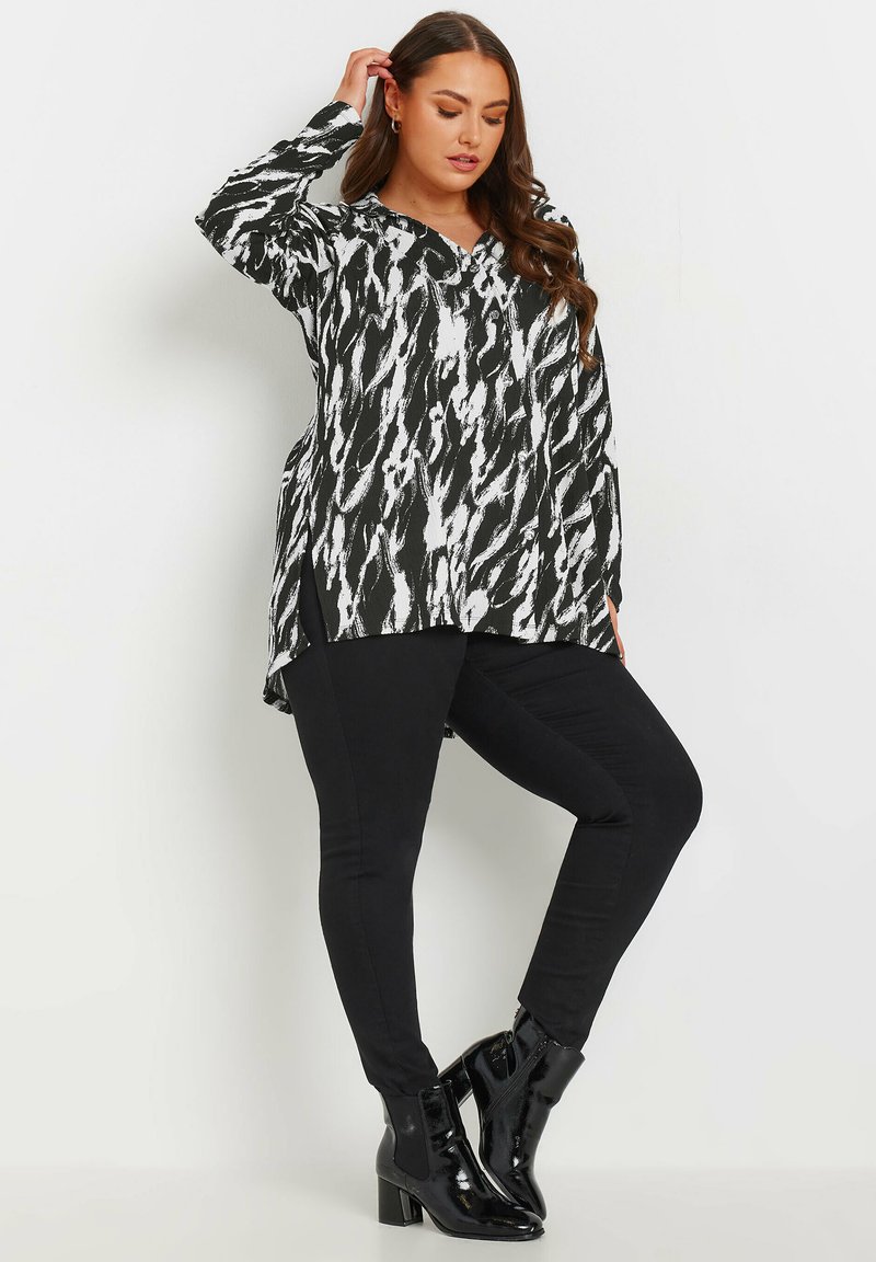 Top lungo a maniche lunghe bianco e nero con scollatura a V, abbinato a pantaloni neri slim-fit e stivaletti neri lucidi.