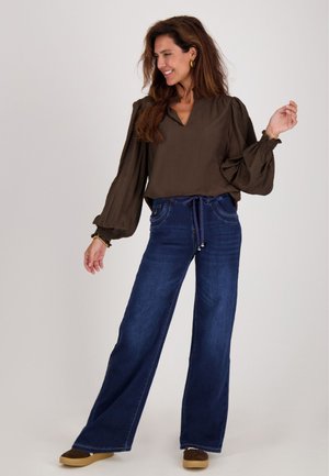 Bruine v-hals blouse met puffmouwen, gecombineerd met donkerblauwe wijde jeans en bruine schoenen, tegen een effen achtergrond.