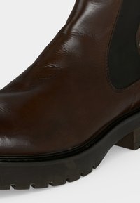 Gros plan d'une botte en cuir marron avec un panneau latéral élastique noir et une épaisse semelle crantée noire sur fond blanc.