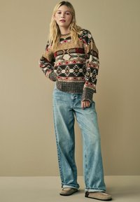 Pull en tricot marron, crème et rouge avec des motifs géométriques. Associé à un jean bleu clair ample et des chaussures de style moccasin beiges.