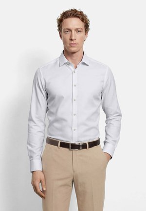 Mann mit lockigem Haar, der ein weißes, geknöpftes Hemd trägt, in beige Hosen gesteckt mit einem dunkelbraunen Gürtel, vor einem schlichten Hintergrund stehend.