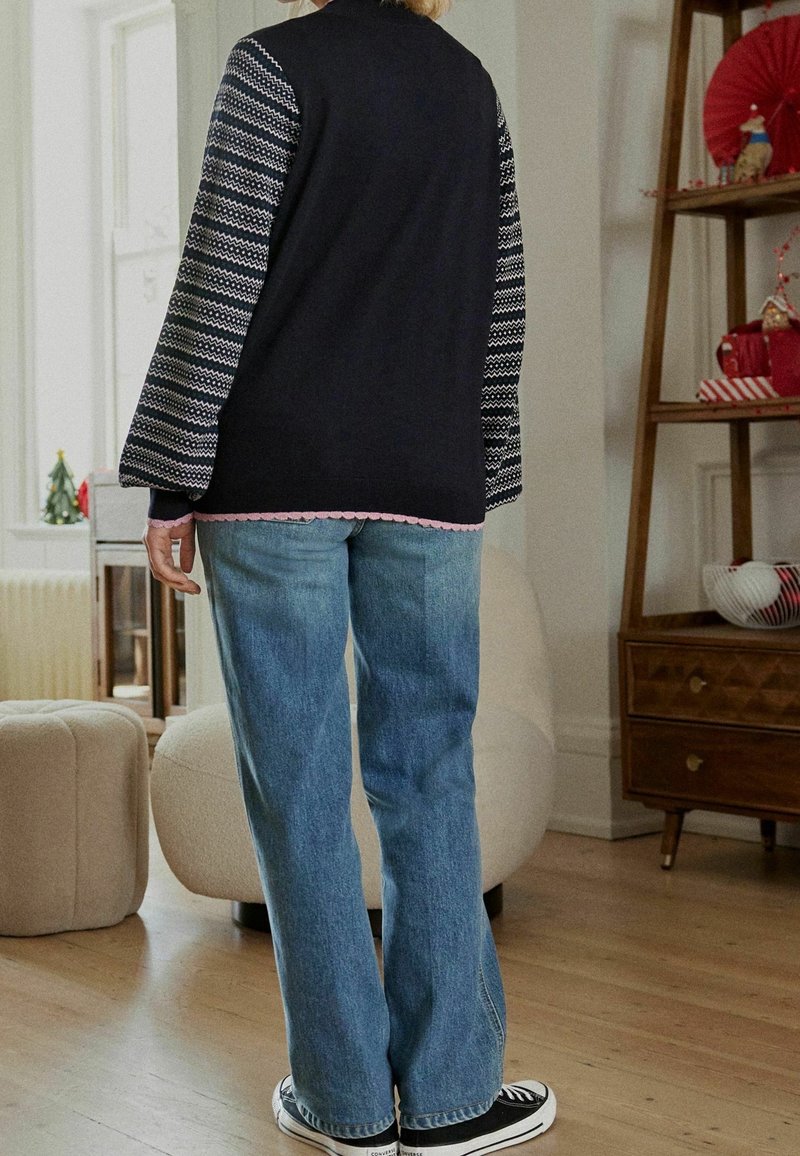 Schwarzer Cardigan mit gemusterten Ärmeln, rosa gewelltem Saum, kombiniert mit hellblauen Jeans mit geradem Bein und schwarzen Sneakern. Holzmöbel im Hintergrund.