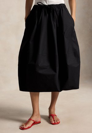 Γυναίκα που φοράει μαύρη φούστα μέχρι το γόνατο με τσέπες και κορδόνι στη μέση, συνδυασμένη με κόκκινα σανδάλια τύπου thong σε γυμνά πόδια.