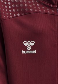 Top deportivo burdeos con una tela texturizada de brillo suave y un patrón circular cerca del escote. Logo "hummel" blanco en el centro.