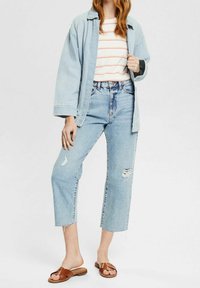 Lichtblauwe denim jeans met gescheurde details, gecombineerd met een wit en oranje gestreept shirt en een lichtblauwe oversized jas. Bruine instapsandalen.