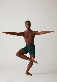 Model in groene shorts, die een balanspose op één voet uitvoert met uitgestrekte armen, waarmee een mooi uitgelijnd lichaam wordt getoond tegen een neutrale achtergrond.