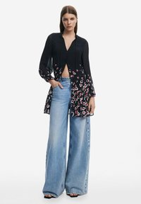 Schwarze Bluse mit Knopfleiste und blumenförmigem Saum, langen Ärmeln und V-Ausschnitt, kombiniert mit weit geschnittenen hellblauen Jeans. Gewebter Stoff.