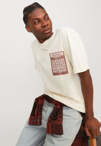 T-shirt en coton couleur crème avec un motif graphique marron sur la poitrine. Associé à un jean en denim bleu clair et une chemise à carreaux rouges nouée à la taille.