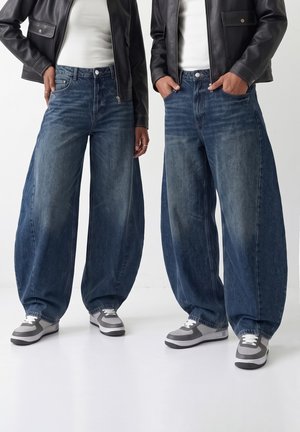 Deux personnes portant des jeans bleus à jambes larges, des baskets blanches, des hauts blancs et des vestes en cuir noires, debout avec les mains dans les poches et sur les côtés.