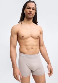 Άνδρας με μακριά ντραντλόκ, χωρίς πουκάμισο, που φοράει ανοιχτό μπεζ μποξεράκια Calvin Klein, στέκεται μπροστά σε απλό φωτεινό φόντο.