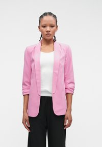 Svetloružový blazer s klopovým golierom, dlhými rukávmi a voľným strihom, kombinovaný s bielym topom a čiernymi nohavicami. Hladká textúra látky.