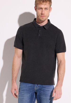 Homme aux cheveux blonds courts portant un polo noir à manches courtes et un jean bleu, debout devant un fond blanc.