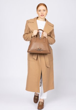 Brown leather handbag with a structured design, dual top handles, logo detail, and zip closure. Presented with a camel coat over white attire.