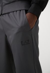Grå atletiske bukser lavet af tekstureret stof, med et diskret EA7-logo og sidelommer, samt en elastisk talje for en sikker pasform.
