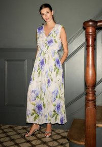 Virágmintás maxi ruha fehér alapon kék és zöld mintákkal, V-nyakú, ujjatlan, derékpánttal. A modellen ezüst színű magassarkú van.