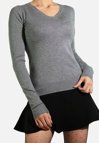 Pull gris à manches longues et col en v en tissu tricoté lisse, avec des poignets et un ourlet côtelés. Associé à une jupe noire évasée et des collants à pois.