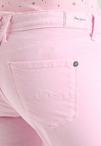 Detail blízkého záběru na růžové džíny, kapsa se šitým designem a značkovou etiketou "Pepe Jeans London" na pasu.